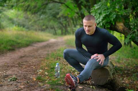 Laktatdiagnostik: Junger Mann im Jogging-Outfit sitzt auf einem Baumstamm im Park und hält seinen schmerzenden Oberschenkel