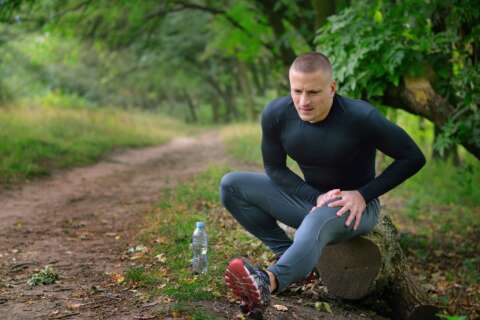 Laktatdiagnostik: Junger Mann im Jogging-Outfit sitzt auf einem Baumstamm im Park und hält seinen schmerzenden Oberschenkel